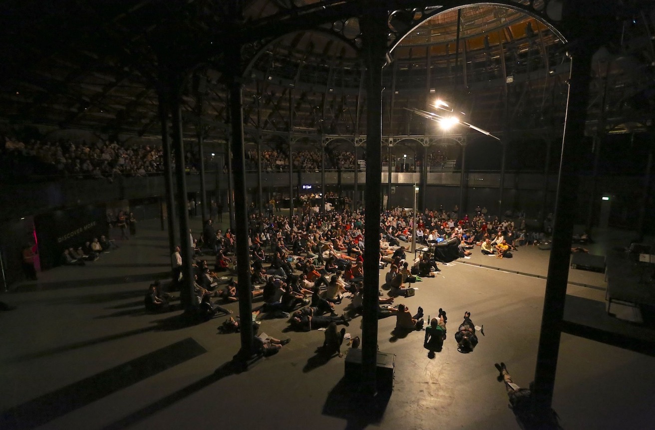 Audience seated and lying on the floor of a large industrial venue